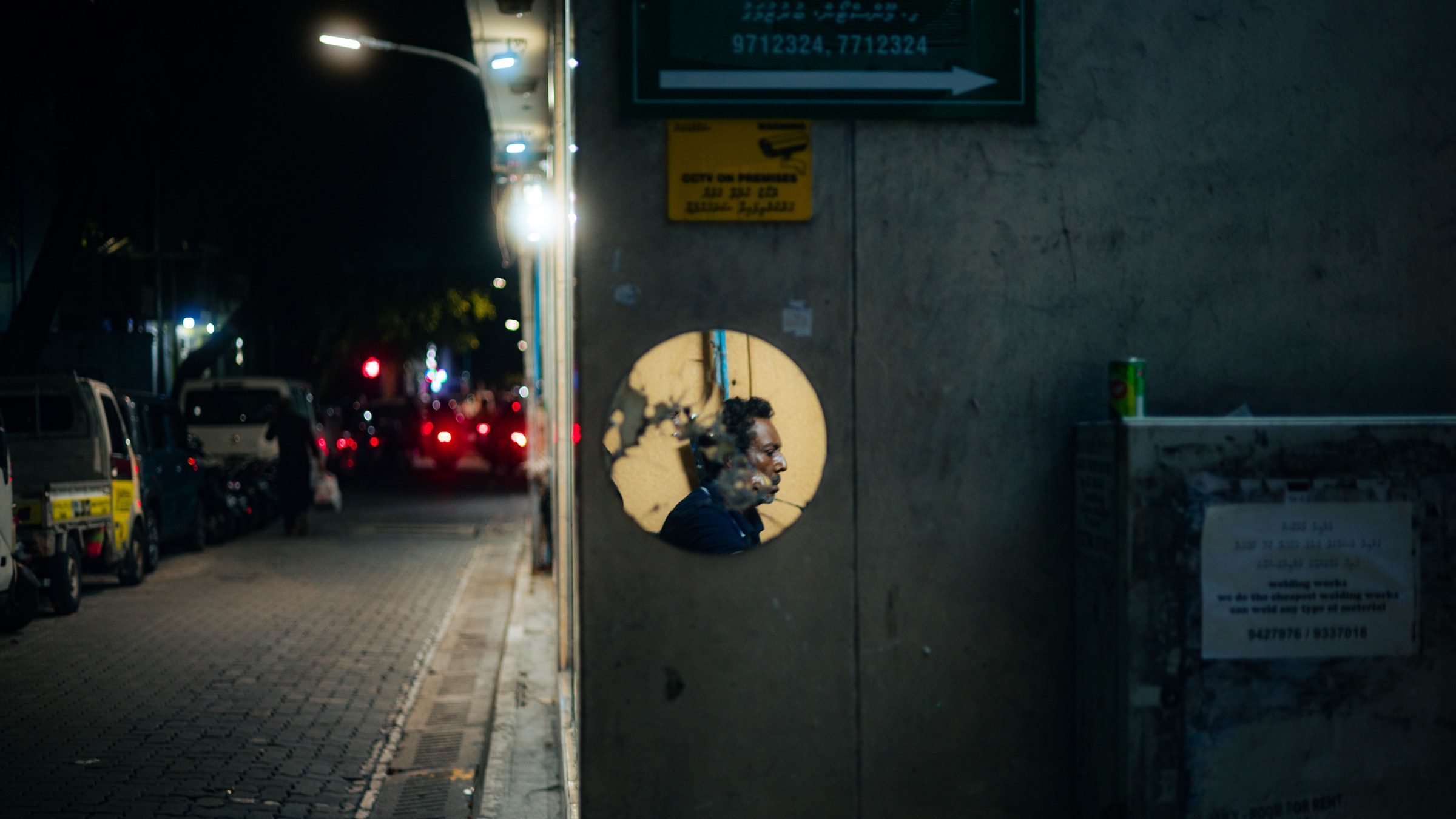 Man's face visible through a circular hole in a wall at night, traffic bokeh in the distance