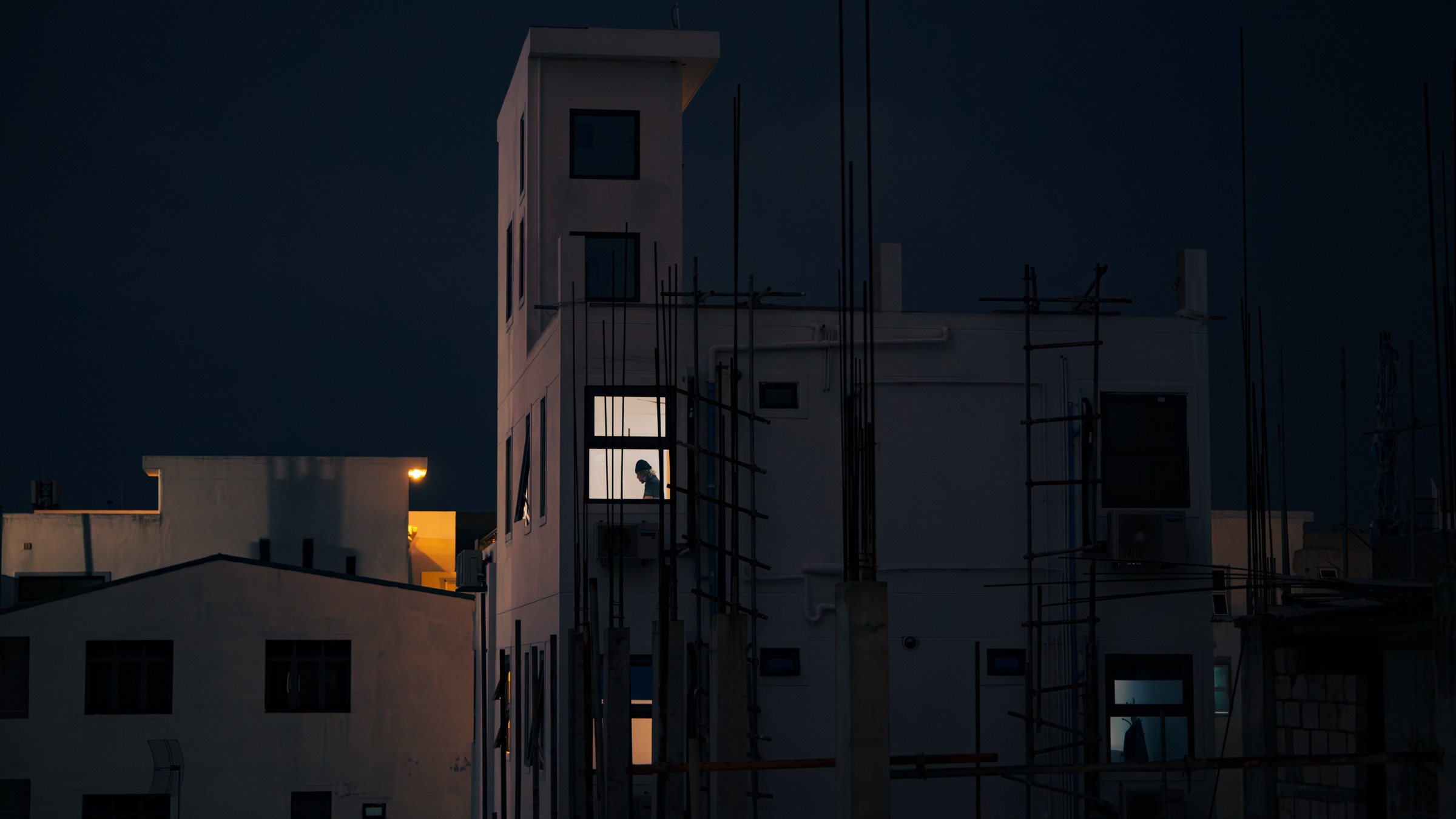 Lone figure silhouetted in a single lit window across dark apartment rooftops at night