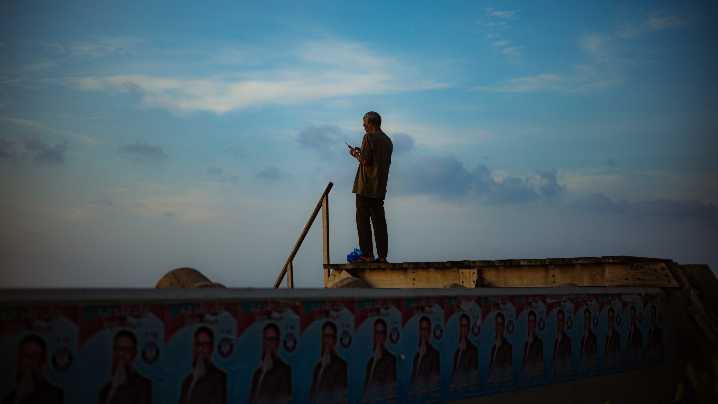 Man standing on a concrete ledge with his phone, election posters along the seawall below
