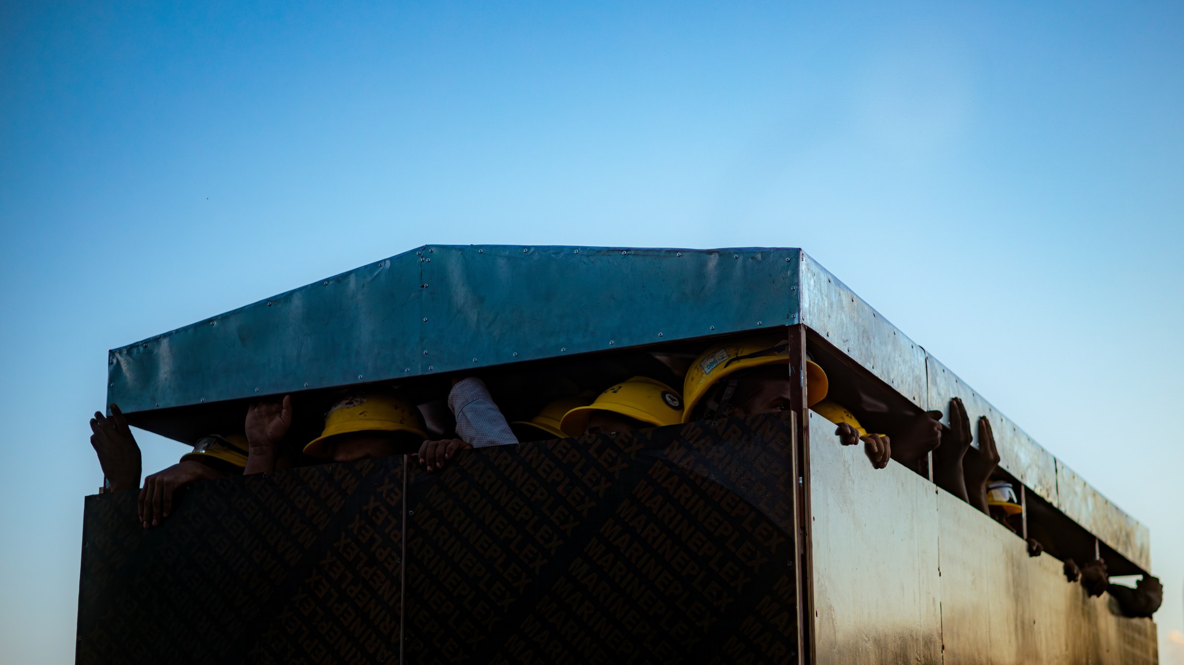 Workers in yellow hard hats crammed under a low metal truck roof against a clear blue sky