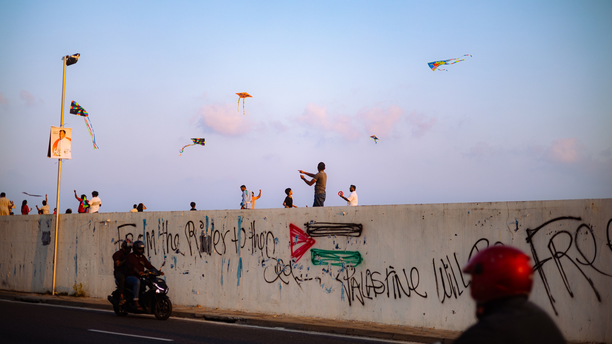 People flying kites above a graffiti-covered seawall, motorcycles passing below at dusk