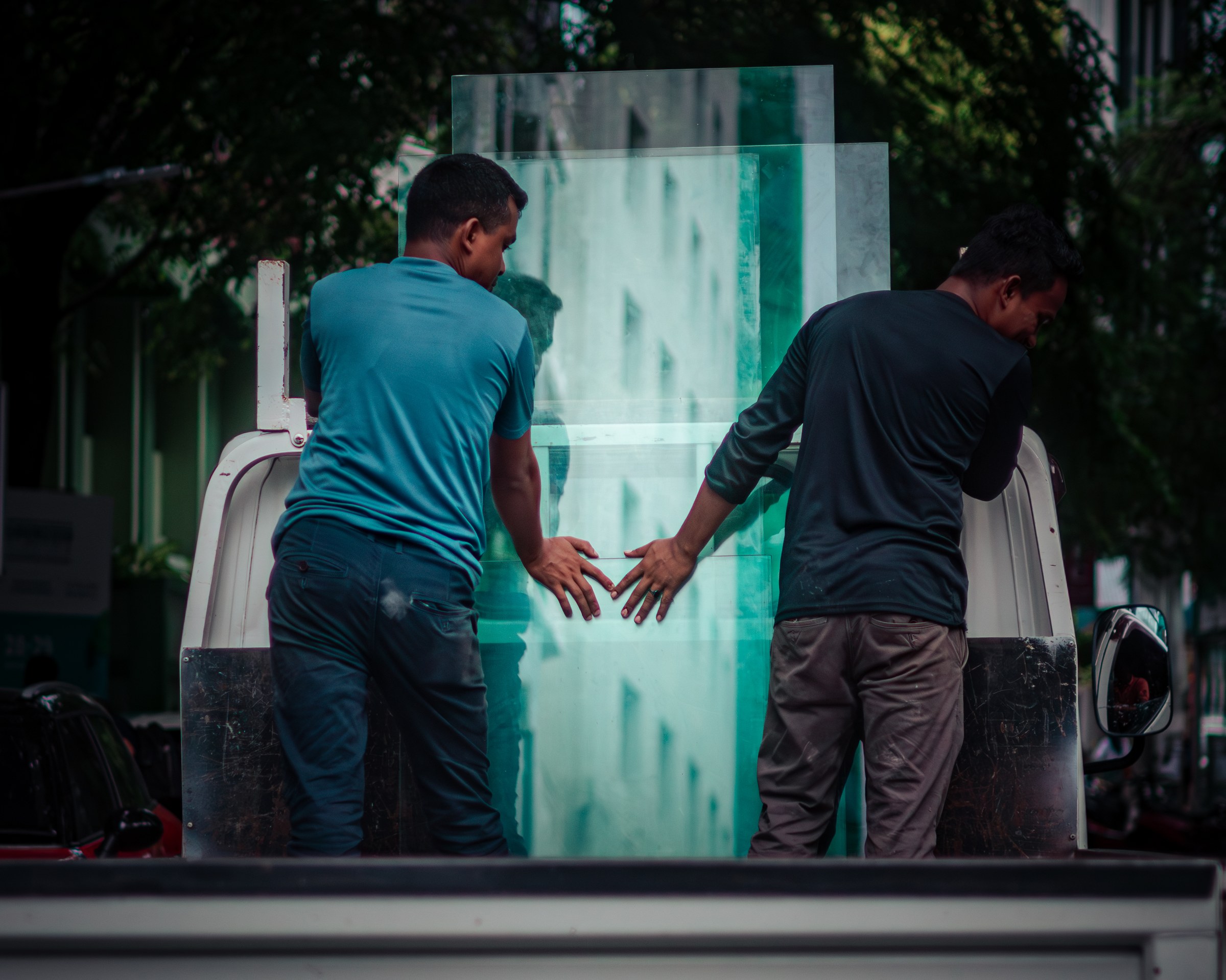 Two workers steadying a large panel of green glass on a truck bed, street trees behind