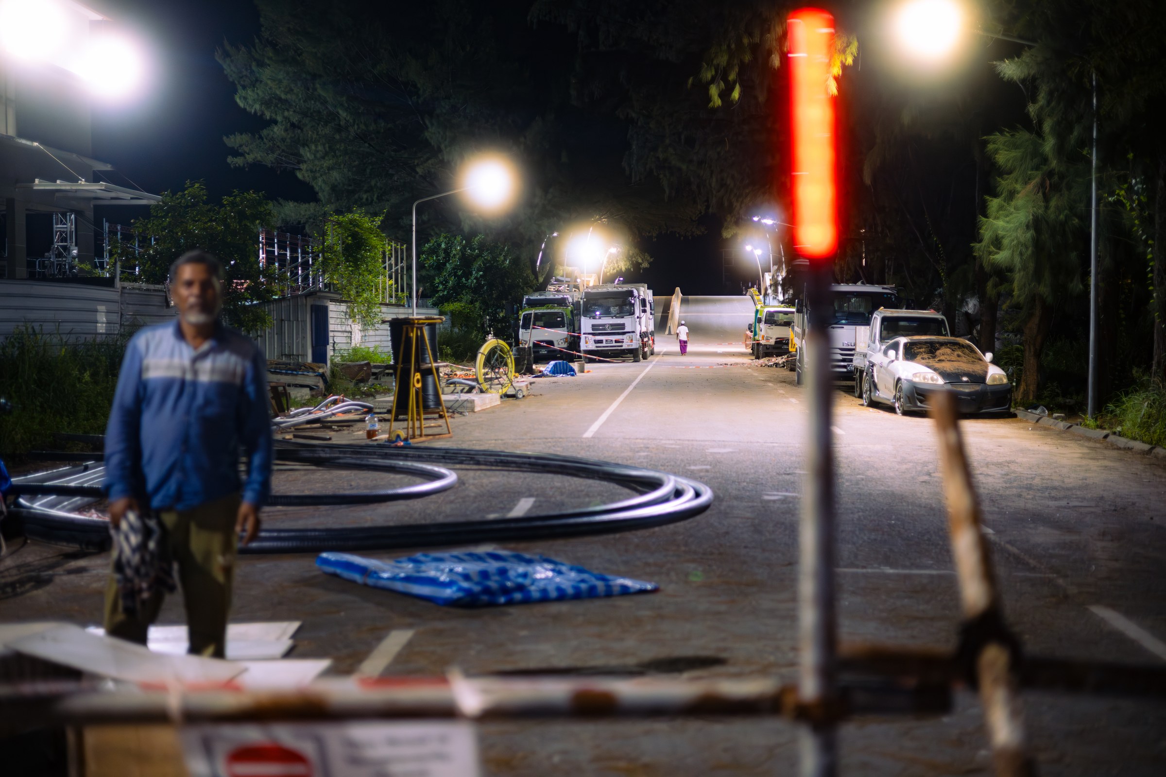 Night road works on a quiet street, construction trucks lit and a lone worker watching
