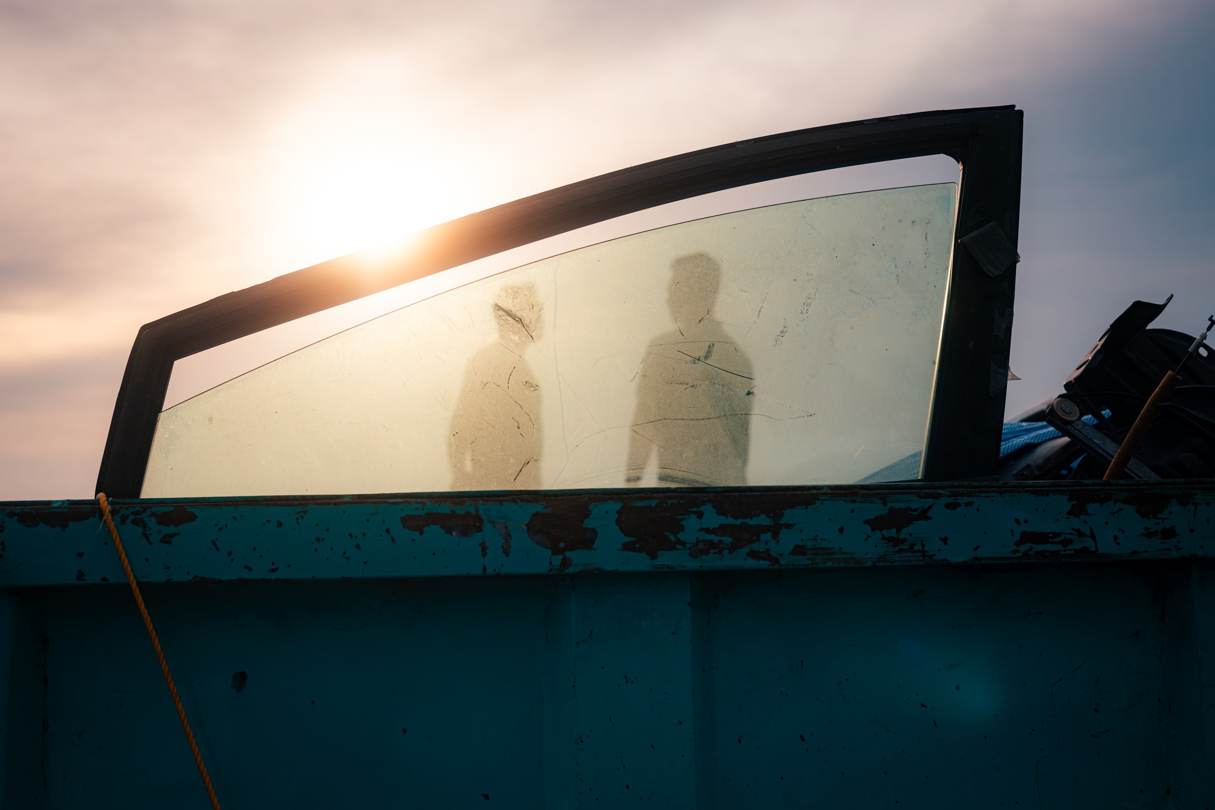 Two shadow silhouettes cast on a scratched car window glass against a golden sunset sky