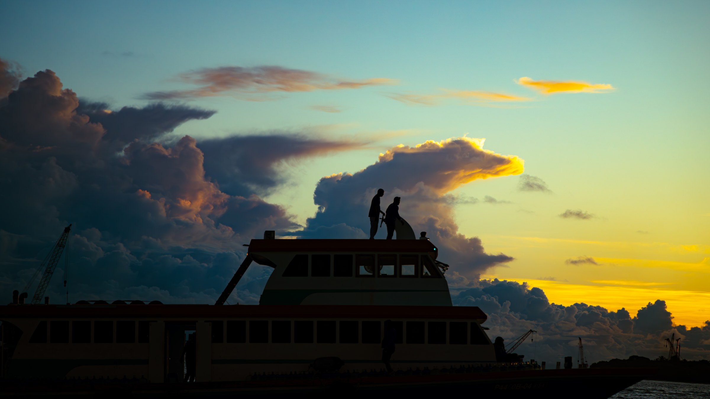 Two workers silhouetted atop a ferry against a dramatic gold and blue sunset sky