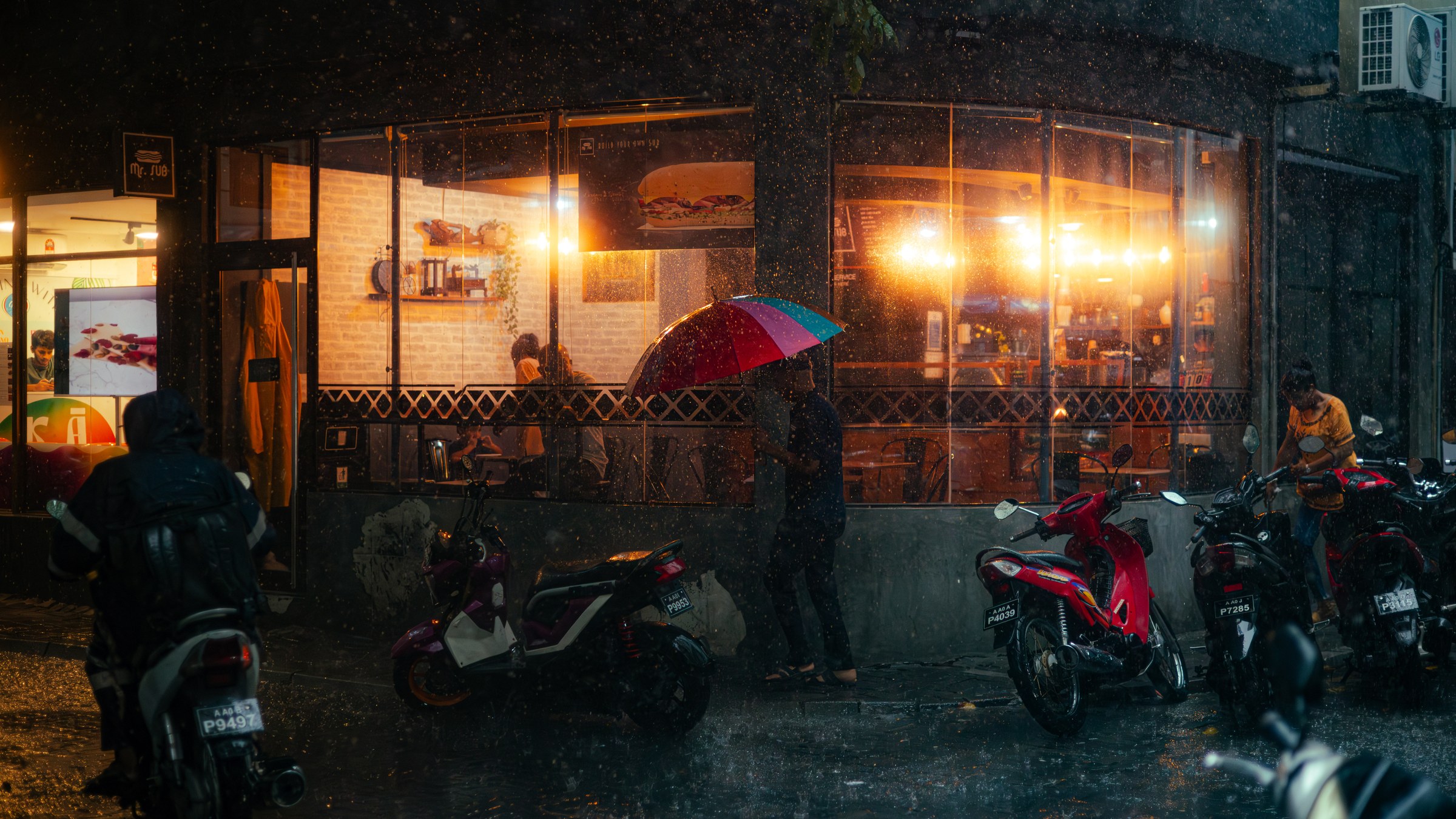 Person sheltering under a rainbow umbrella outside a warm café in the rain