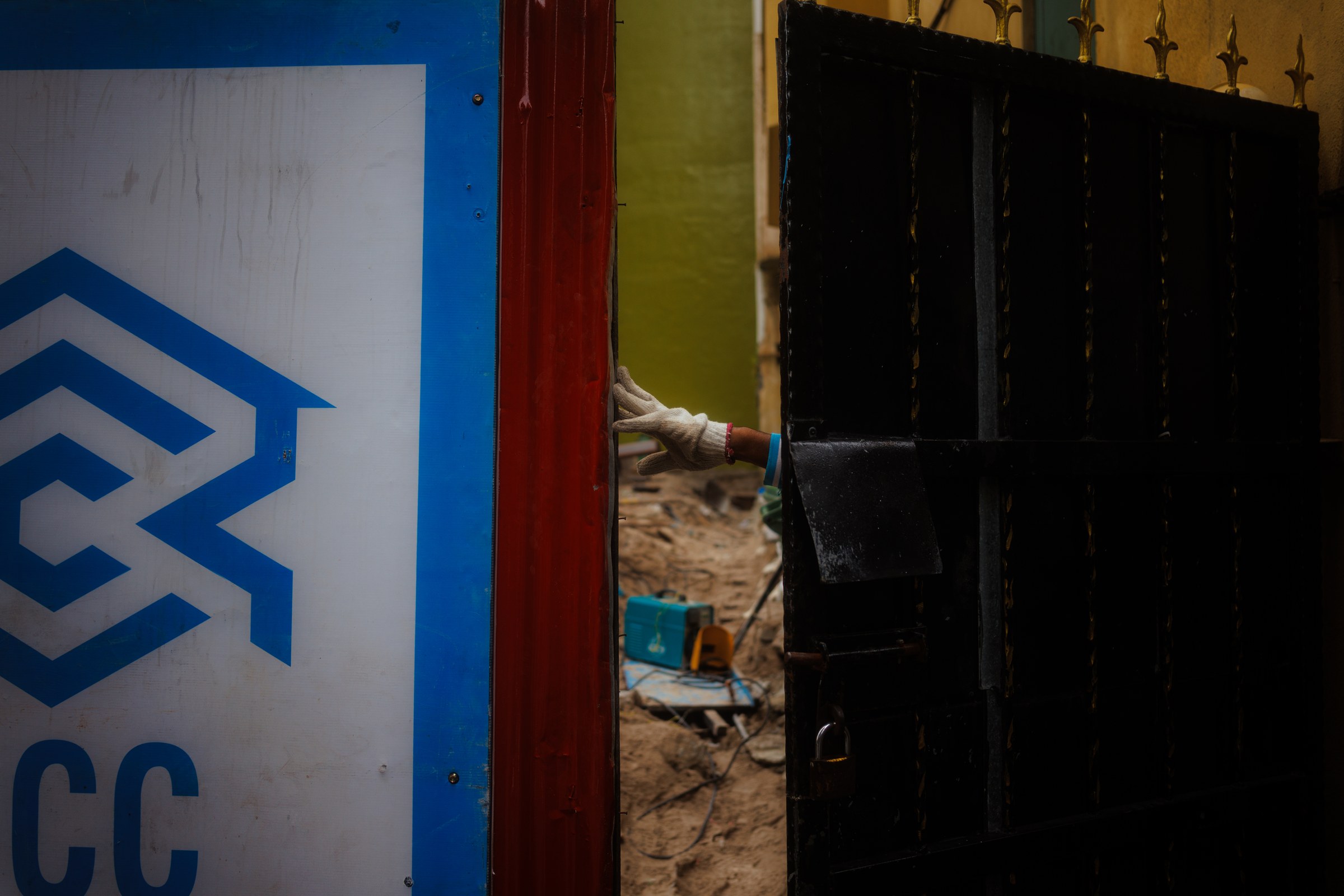 Gloved worker's hand visible through a gap between a construction hoarding and an iron gate