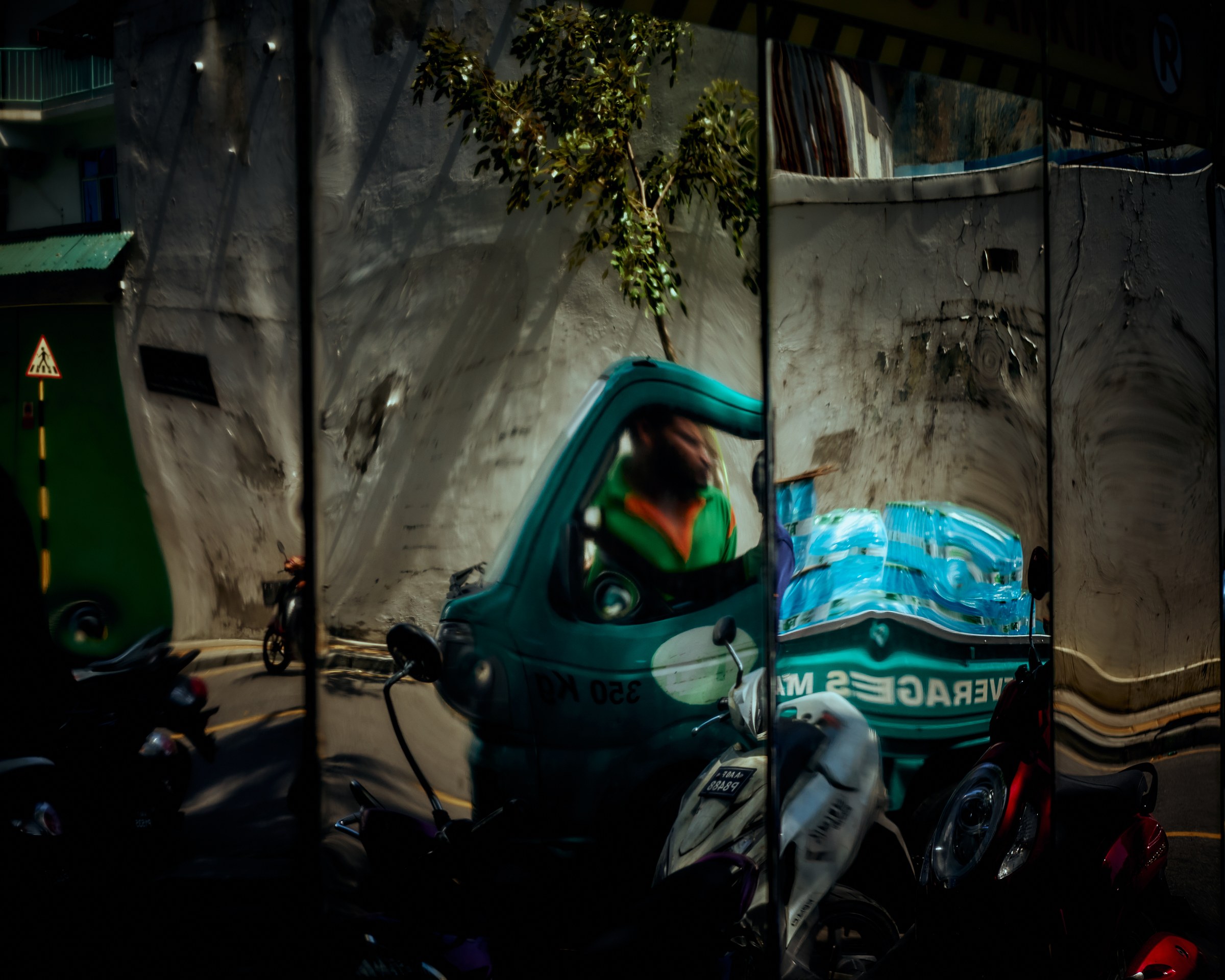 Street scene distorted in a shop window reflection — delivery tuk-tuk and motorcycles on a busy lane