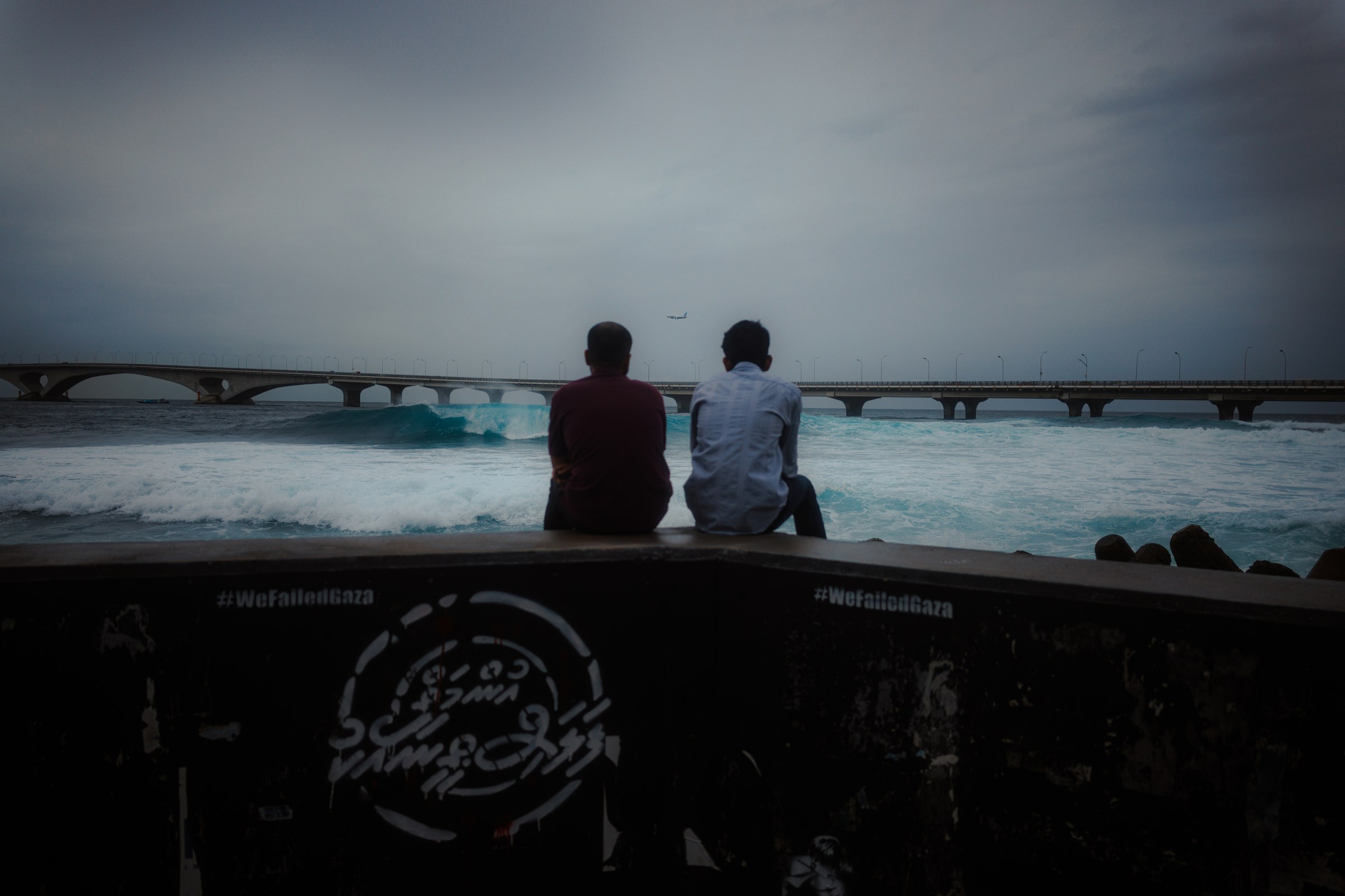 Two men sitting on a seawall, backs to camera, facing the bridge and a choppy grey ocean