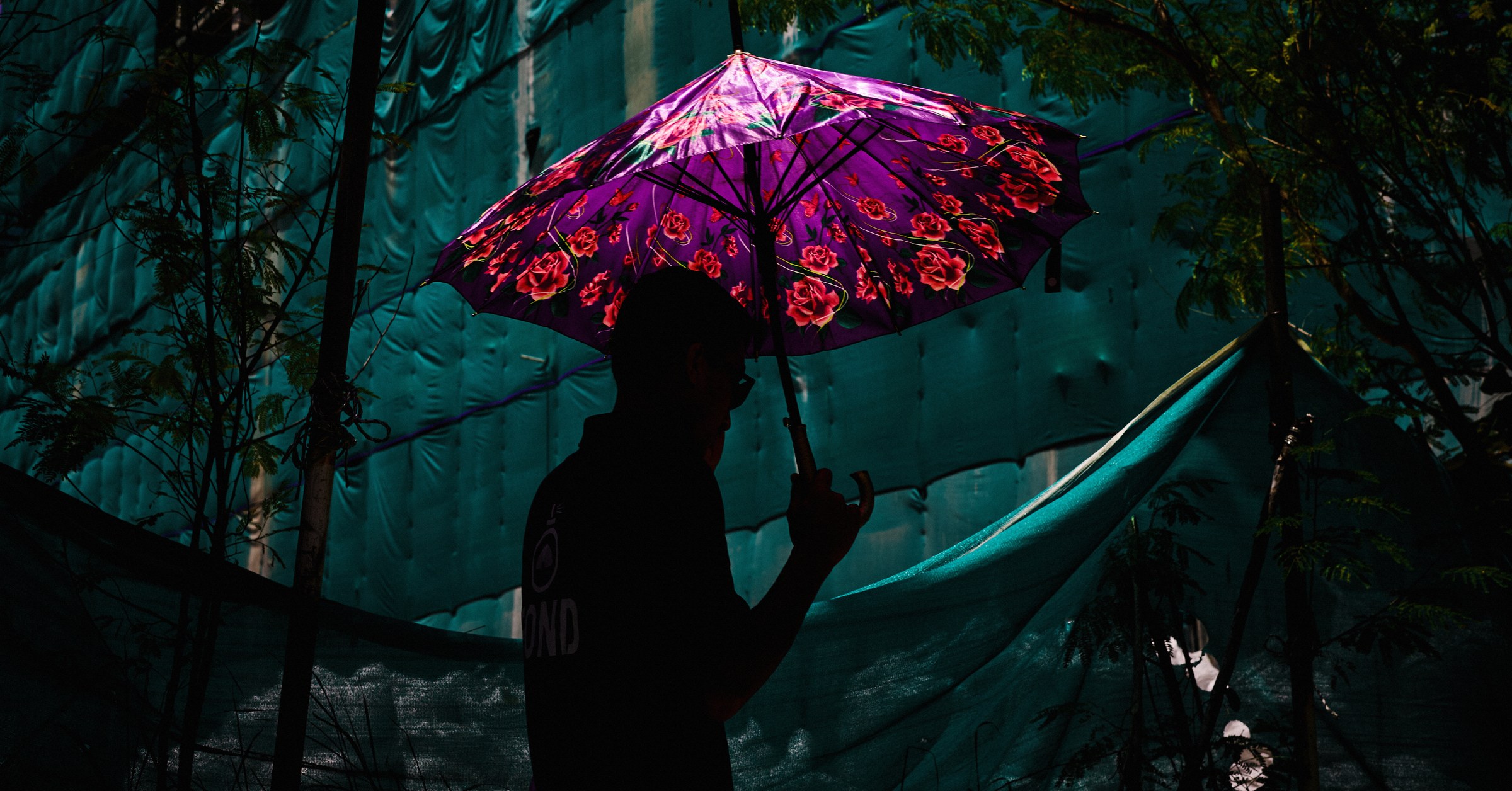 Figure holding a vibrantly lit floral umbrella, silhouetted against dark green scaffolding nets at night