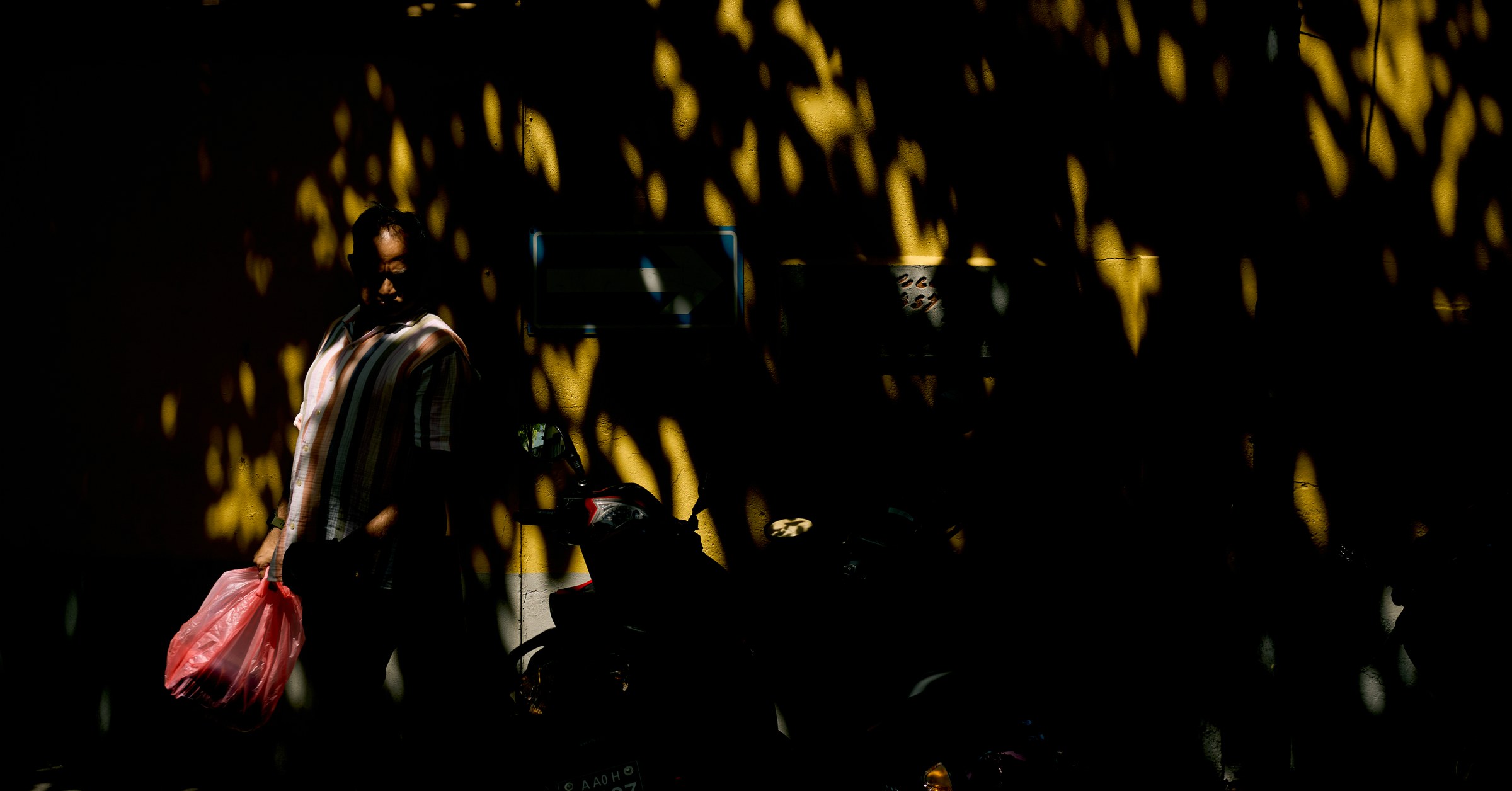 Man carrying a pink plastic bag through dappled golden light falling on a dark street