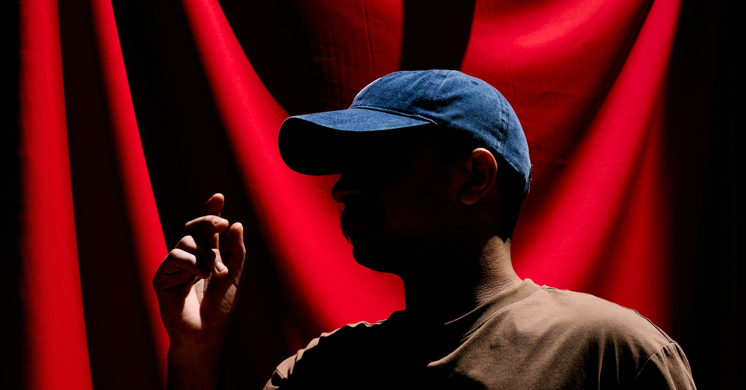 Man in a blue cap, face in shadow, profiled against deep red draped fabric