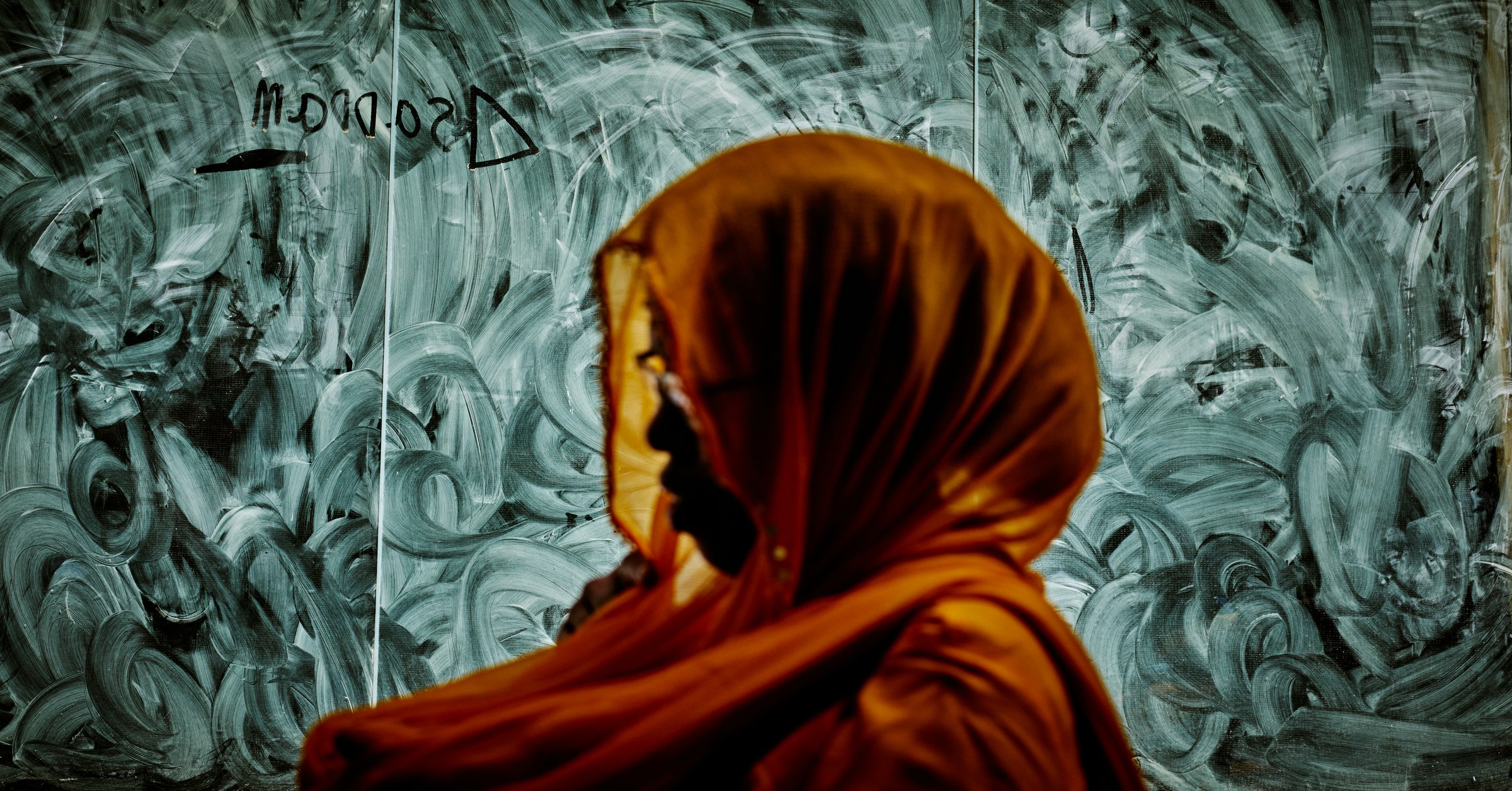 Woman in orange hijab in sharp profile against a grey swirling painted wall