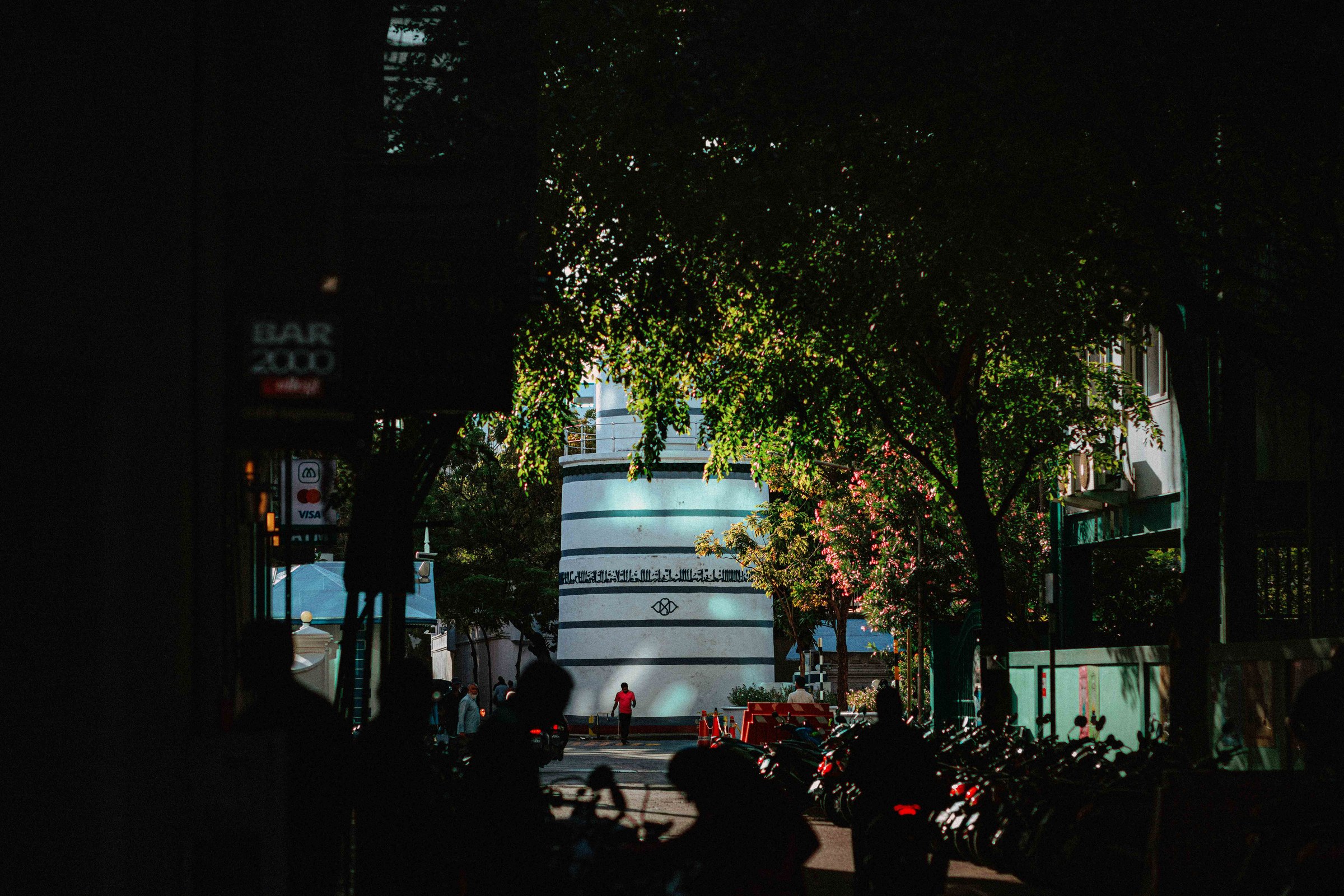 Street framed by dark trees, lone figure in red near a glowing circular building in the distance