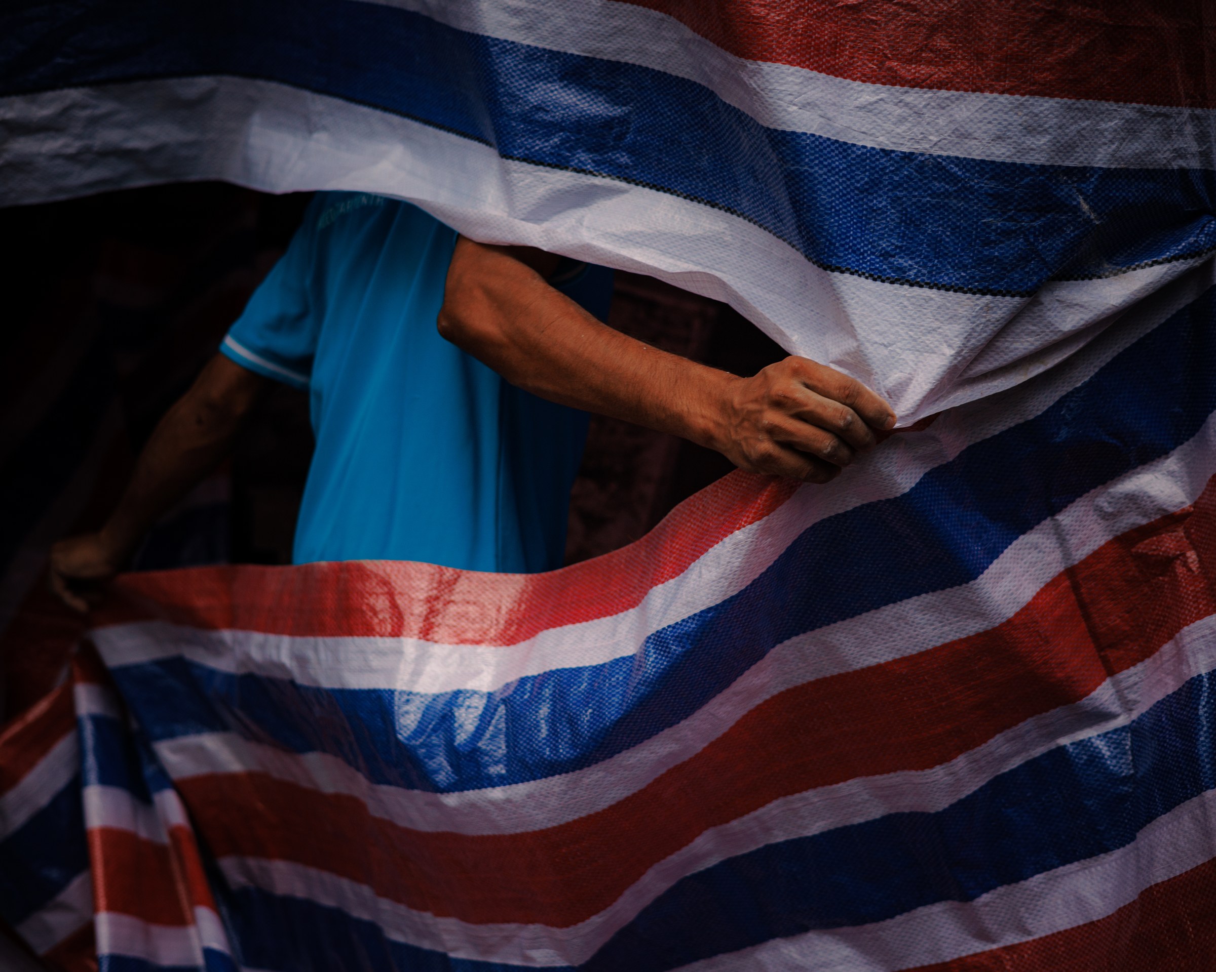 Worker's hand gripping striped red, white and blue tarpaulin, blue shirt visible behind