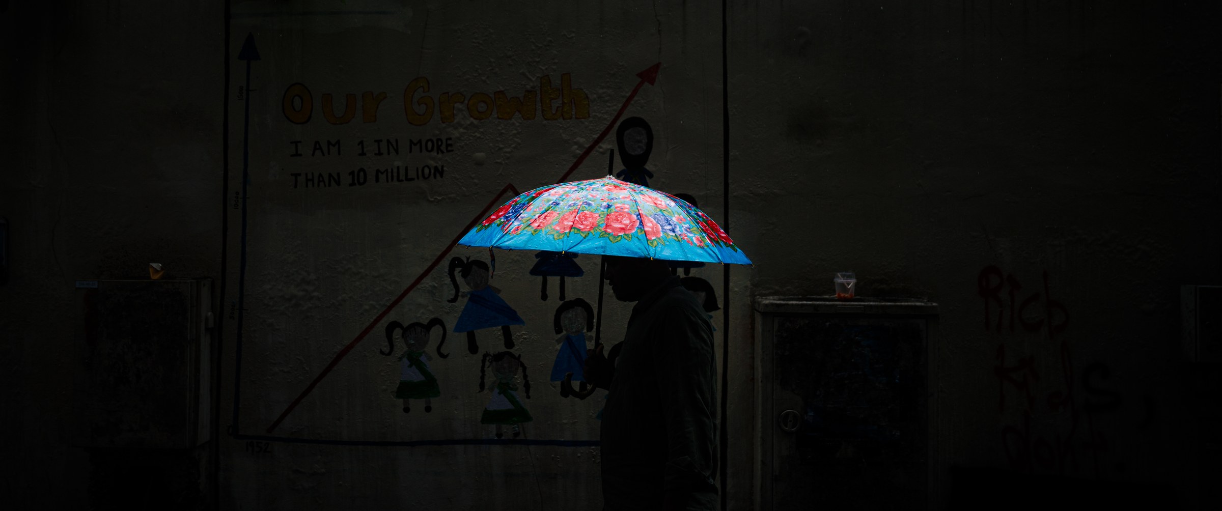 Silhouetted figure with a floral umbrella passing a painted children's mural on a dark rainy street