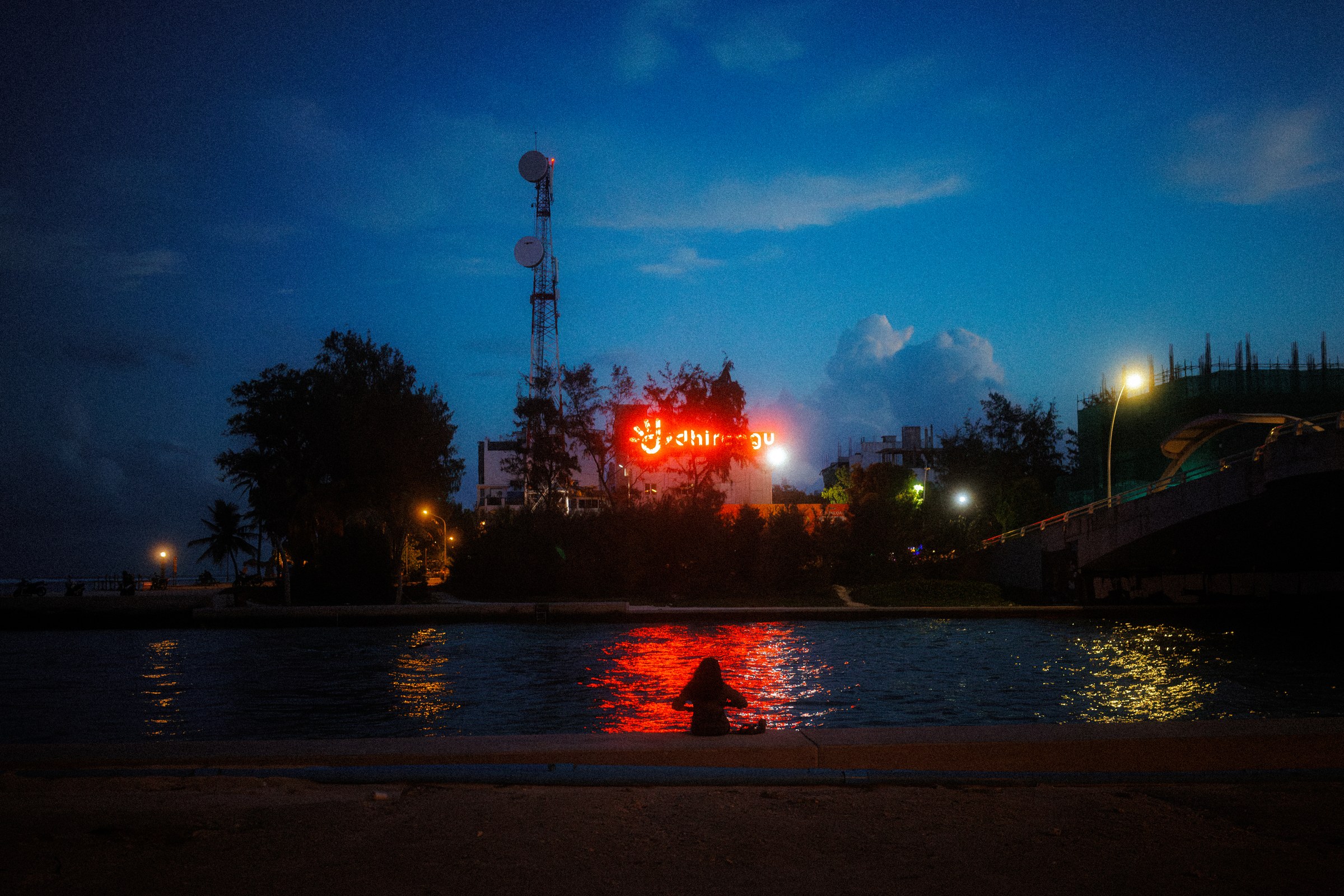 Lone figure sitting by the waterfront at night, red billboard light reflecting on the surface