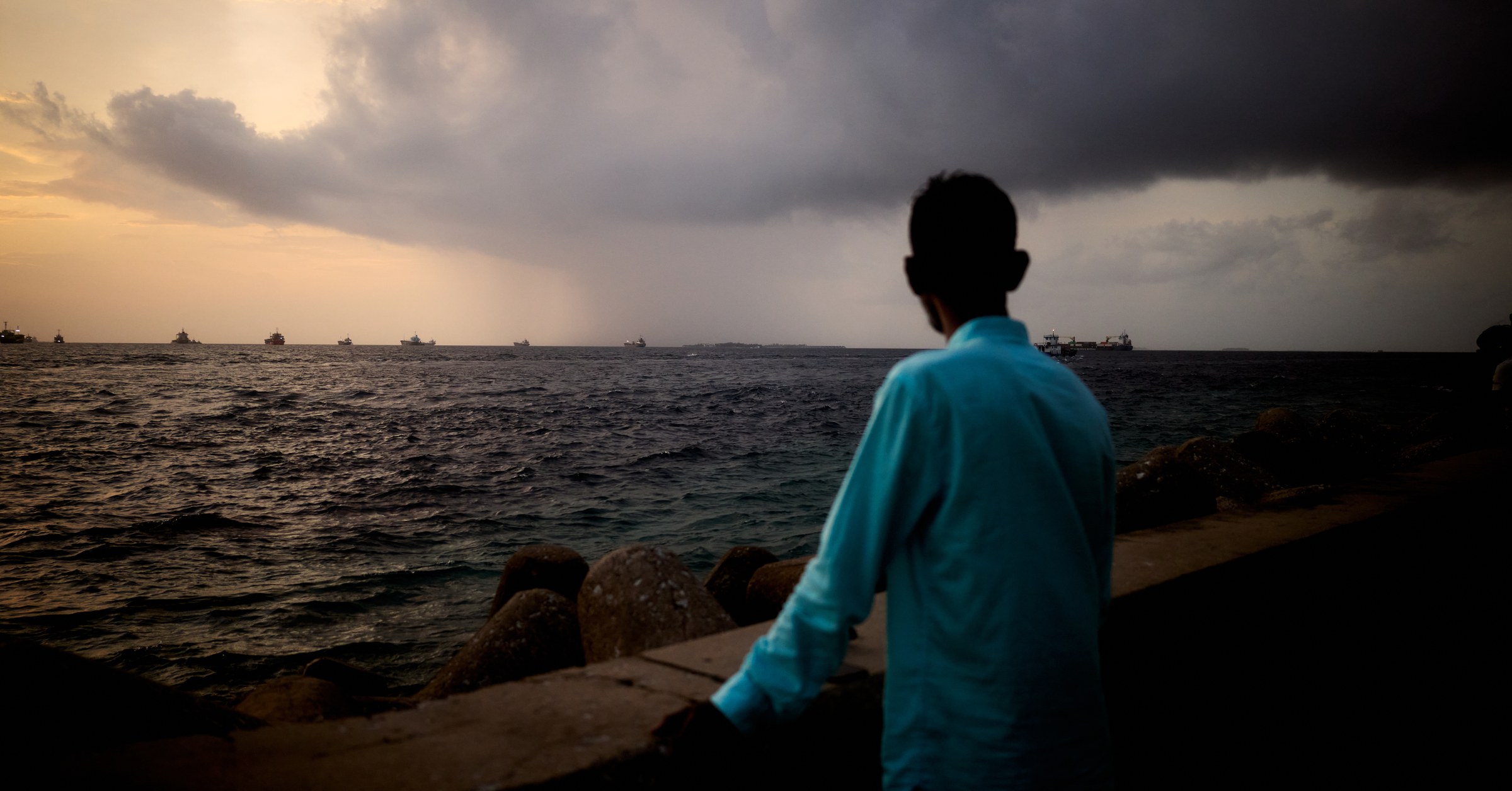 Man in a light blue shirt watching a storm roll in from the sea, ships on the horizon at dusk